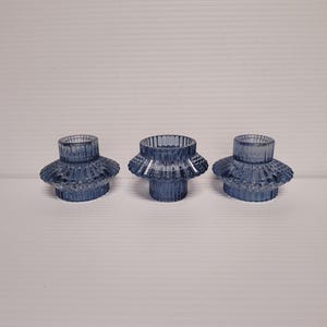 May include: Three blue glass candle holders. Each holder has a cylindrical base and a ruffled, flared top. The glass has a textured, ribbed design. The candle holders are arranged in a row against a plain background.