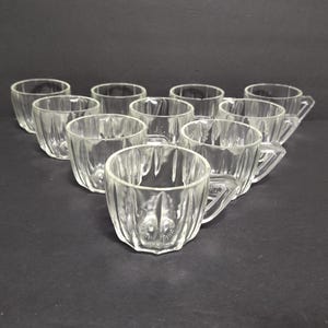 May include: A collection of clear glass teacups arranged on a dark surface. Each cup features a faceted design and a small handle. The cups are empty and appear to be vintage.