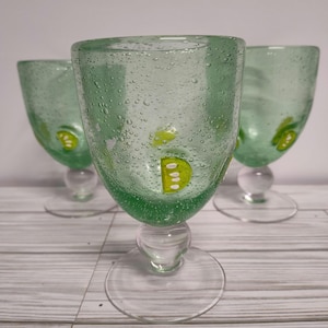 May include: Three green glass wine glasses with a textured surface and yellow and white designs. The glasses have a round base and a stem.
