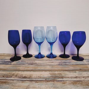 May include: Six wine glasses in varying sizes, arranged in a row. The glasses alternate between a deep blue and a lighter blue hue. The stems and bases are dark blue, while the bowls vary in color.