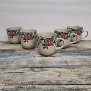 May include: A set of six off-white ceramic mugs. Each mug features a red apple design with green leaves and a checkered border. The mugs have a rounded shape and a handle. The background is a light blue and brown wooden surface.