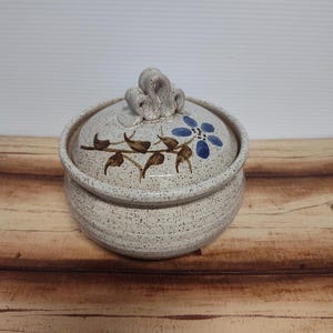 May include: A speckled ceramic jar with a lid. The lid features a blue floral design and a decorative handle. Brown leaves and stems are painted around the flower. The jar is sitting on a wooden surface.