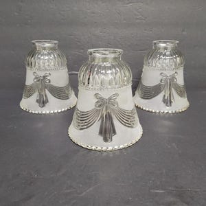 May include: Three vintage glass lamp shades. Each shade has a frosted white body with a silver bow and ribbon design. The top and bottom edges are trimmed with a silver beaded design. The shades are bell-shaped.