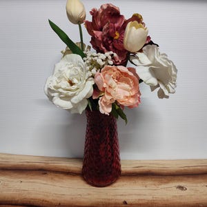 May include: A vibrant floral arrangement in a deep red glass vase. The bouquet features white roses, peach peonies, and burgundy and cream-colored tulips. The vase has a textured diamond pattern.