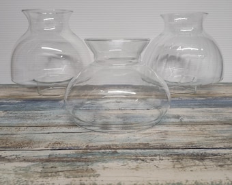 May include: Three clear glass lamp shades with a rounded, bulbous body and a flared top opening. The shades feature a smooth, transparent surface and a circular base designed for attachment to a light fixture, arranged in a group of three.