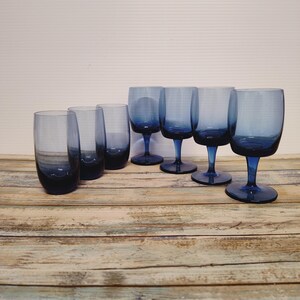 May include: A collection of six blue-tinted drinking glasses. Three are stemless tumblers, and three are stemmed wine glasses. The glasses are arranged on a weathered wooden surface, with a plain white background.