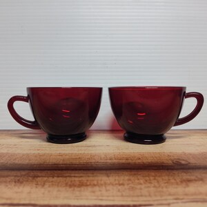 May include: Two ruby red glass teacups with handles. The cups have a rounded bowl shape and sit on a small, dark base. The cups are displayed on a wooden surface against a white background.