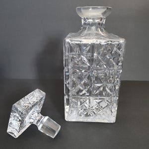 May include: A clear, square-shaped crystal decanter with a matching stopper. The decanter and stopper feature a geometric, diamond-like pattern. The stopper is lying next to the decanter on a dark surface.