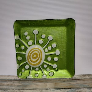 May include: Square green glass decorative plate with a white and gold spiral design in the center. The plate has a sunburst pattern with white dots and lines. The plate is on a wooden surface.