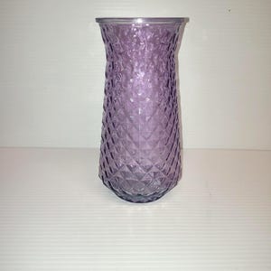May include: A translucent purple glass vase featuring a textured diamond pattern across its exterior surface. The vase has a wide, circular opening at the top that tapers slightly inward before widening toward the base, creating a distinct silhouette.