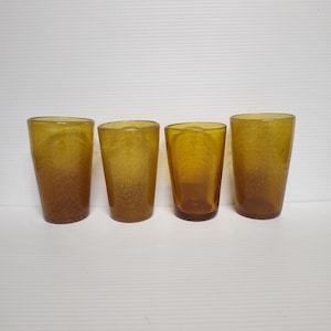 May include: Four amber-colored drinking glasses. The glasses have a textured appearance and a slightly tapered shape. The color gradient transitions from a darker amber at the base to a lighter amber towards the rim. The glasses are arranged in a row.