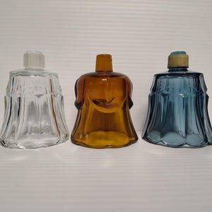 May include: Three glass candle holders in clear, amber, and blue. Each has a bell-shaped design with a flared base and a small opening at the top. The clear one has a ribbed top, while the blue one has a gold-colored top.