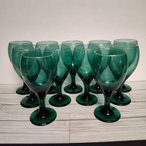 May include: Set of ten green wine glasses with gold rims. The glasses are arranged in a semi-circle on a white wooden surface.