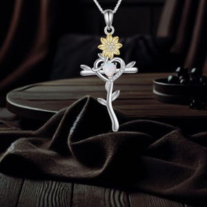 Könnte beinhalten: Eine silberfarbene Halskette mit Anhänger in Kreuzform. Das Kreuz enthält ein herzförmiges Element mit einem weißen Edelstein, einer gelben Blume und einem floralen Stiel. Die Halskette hängt an einer feinen Silberkette.