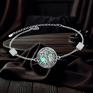 May include: A silver bracelet with a round charm featuring a tree of life design with a green and blue abalone shell inlay. The charm is surrounded by a silver Celtic knot design. The bracelet has two square-shaped accents with clear cubic zirconia stones.