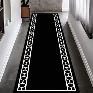 Op de afbeelding: Een lange, rechthoekige zwarte loper met een witte geometrische rand. Het randontwerp heeft een herhalend patroon van verbonden ovalen en cirkels. De loper is geplaatst in een gang.