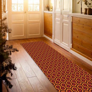 May include: A red and orange patterned runner rug with a geometric design. The rug is placed in a hallway with wooden floors and white doors.