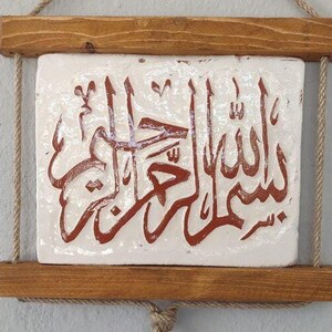 Puede incluir: Un azulejo de cerámica blanca con caligrafía árabe marrón sobre él, enmarcado con dos listones de madera y colgado de una cuerda con un borla.