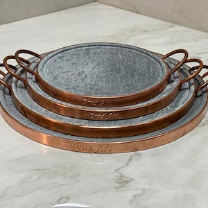 May include: Set of three round, gray stone hot plates with copper rims and handles. The plates are nested inside each other and have the words "Bras'Art" engraved on the rims.