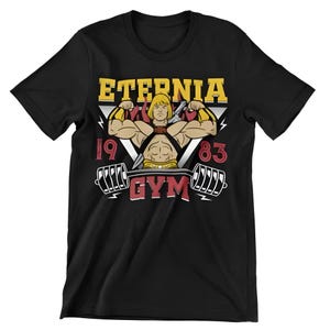 Könnte beinhalten: Schwarzes T-Shirt mit einer Grafik eines muskulösen Cartoon-Charakters und dem Text "ETERNIA GYM 1983". Das Design beinhaltet eine Langhantel und Blitze, mit einem gelb-roten Farbschema.