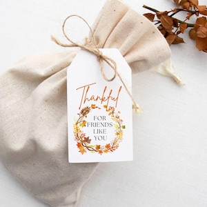 May include: A white gift bag with a brown twine bow and a gift tag that says "Thankful for friends like you" with a wreath of fall leaves and flowers.