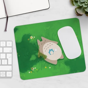 Puede incluir: Una alfombrilla de rat&oacute;n verde con un personaje de dibujos animados, Totoro, tumbado en una cama de hierba. Un rat&oacute;n de ordenador blanco est&aacute; en el lado derecho de la alfombrilla.