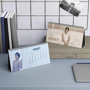May include: A desk calendar featuring a photograph of a person in a light-colored outfit and the year 2027 on the cover, alongside an open page for August displaying a calendar grid and a portrait of the same person in a patterned jacket.