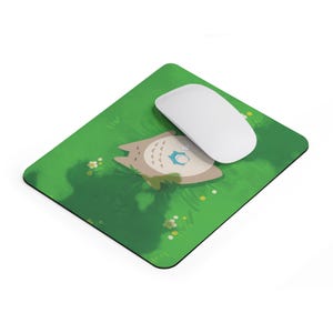 Puede incluir: Una alfombrilla de rat&oacute;n verde con una imagen de dibujos animados de una criatura gris y marr&oacute;n con un p&aacute;jaro azul en la cabeza. La criatura est&aacute; tumbada en una cama de hierba verde con flores blancas. Un rat&oacute;n de ordenador blanco est&aacute; en la alfombrilla.