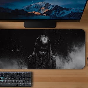 May include: Black desk mat with a grayscale illustration of a person with long hair and a dark background. A black keyboard, mouse, and monitor are on the wooden desk. Two potted plants are also on the desk.
