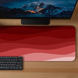 May include: A red and pink gradient mousepad with a black keyboard and mouse. The mousepad has a wave-like design. A computer monitor and two potted plants are also visible on the wooden desk.