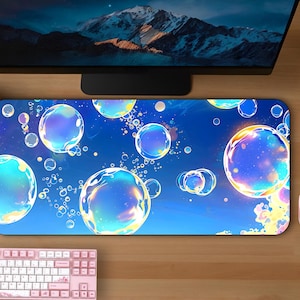 May include: A blue mousepad with a bubble design, a pink mouse, and a pink keyboard are on a wooden desk. The mousepad features large, colorful bubbles against a blue background. A computer monitor and two potted plants are also visible.