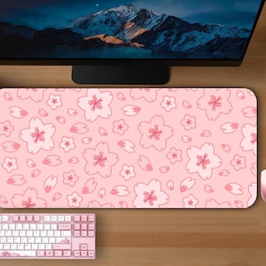 May include: A pink mouse mat featuring a pattern of white and pink cherry blossom flowers and petals. A matching pink mouse and keyboard are also visible. The setup is on a wooden desk, with a computer monitor and plants in the background.