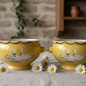 Peut inclure: Deux bols en céramique jaune avec des anses latérales, ornés d'un motif peint représentant deux oies blanches portant des rubans bleus. Les bols présentent un bord festonné noir et une base sur pied, entourés de fleurs de marguerites blanches.