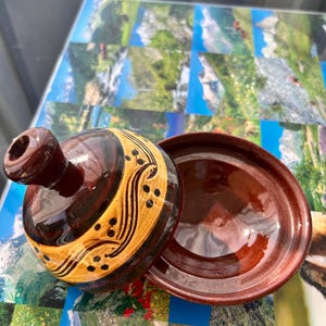 Peut inclure: Un tajine en céramique vernissée composé d'une base ronde profonde et d'un couvercle conique. Le couvercle présente un sommet brun foncé et une bande beige ornée de lignes tourbillonnantes et de petits points peints à la main en noir.