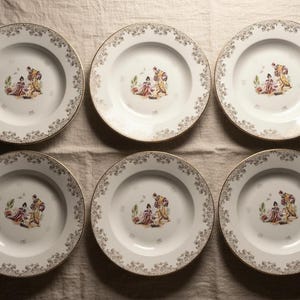 Può includere: Sei piatti in porcellana bianca con bordo dorato e motivo floreale decorativo. Ogni piatto presenta un'immagine centrale di figure in abiti d'epoca. I piatti sono disposti su un panno di colore neutro.