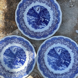 Peut inclure: Trois assiettes rondes en céramique avec une base blanche et un motif bleu de type transfert. Le centre de chaque assiette présente un paysage avec des arbres et un bâtiment, tandis que le bord festonné est orné d'une bordure florale détaillée.