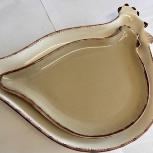 Könnte beinhalten: Zwei Keramik-Servierplatten in Form eines Huhns. Die obere Platte ist hellbraun, die untere weiß. Beide haben einen braunen Rand. Die Platten sind ineinander verschachtelt.