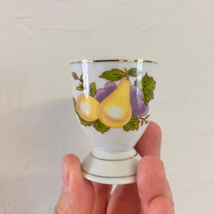Könnte beinhalten: Ein Eierbecher aus weißem Porzellan mit einer dekorativen Illustration einer gelben Birne und violetten Weintrauben mit grünen Blättern. Der Rand und der Fuß des Bechers sind mit einem dünnen Goldrand verziert, was für ein elegantes Finish sorgt.