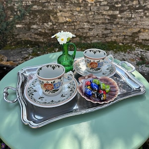 Peut inclure: Un plateau argenté présente deux tasses à thé et soucoupes fleuries, un petit vase vert avec des fleurs blanches et une coquille Saint-Jacques remplie de bonbons colorés. Les tasses ont des motifs floraux orange, bleus et jaunes.