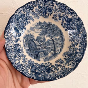 Peut inclure: Un petit bol rond en céramique à bord festonné, orné d'un motif détaillé en bleu et blanc. Le centre représente un paysage rural avec un moulin à eau et des arbres, tandis que le bord est décoré de motifs floraux complexes et détaillés.