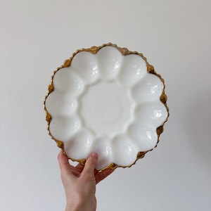 Pode incluir: Um prato branco, com bordas onduladas, para servir ovos com uma borda dourada. O prato tem 12 compartimentos individuais para ovos e um poço central. O prato é segurado por uma mão contra um fundo branco liso.