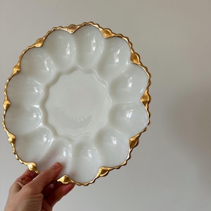 Peut inclure: Plat à œufs en céramique blanche avec un bord festonné doré. Le plat comporte une zone circulaire centrale entourée de 12 empreintes en forme d'œuf. La bordure dorée ajoute une touche d'élégance à ce plat de service.