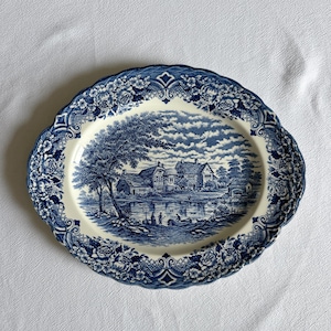 May include: An oval ceramic platter featuring a blue and white transferware pattern. The center depicts a pastoral landscape with houses, trees, and figures by a river. The scalloped rim is decorated with a detailed floral and lattice border.