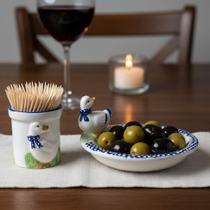 Peut inclure: Une table avec un verre de vin, une bougie allumée et un petit bol d'olives. Un porte-cure-dents en céramique et un canard décoratif sont également présents. Le porte-cure-dents et le canard ont un motif bleu et blanc.