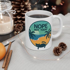Könnte beinhalten: Weiße Keramik-Kaffeetasse mit einer blauen, grünen und gelben Illustration einer Strandszene mit Palmen und einer Katze. Der Text "NOPE NOT TODAY" ist auf die Tasse gedruckt.