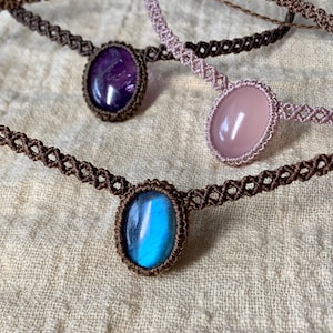 Puede incluir: Tres collares de macramé hechos a mano con colgantes de piedras preciosas de diferentes colores. Los collares son marrones y presentan una amatista morada, un cuarzo rosa rosa y una labradorita azul.