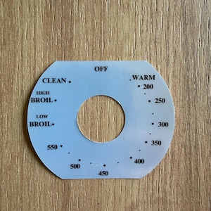 Puede incluir: Un dial de control de horno azul claro con un agujero central. El dial presenta configuraciones de temperatura de 93 a 288 grados Celsius, junto con las configuraciones "OFF", "WARM", "CLEAN" y "BROIL". El dial está sobre un fondo de madera.