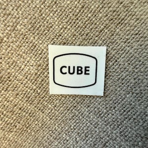 Pode incluir: Uma pequena etiqueta branca com a palavra "CUBE" em letras pretas em negrito. O texto está centrado dentro de uma borda retangular arredondada preta. A etiqueta está sobre um fundo de tecido bege texturizado.
