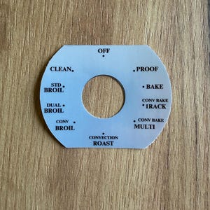 Può includere: Un quadrante di controllo del forno bianco con testo nero. Il quadrante ha un foro centrale e presenta impostazioni come "OFF", "CLEAN", "BAKE" e "CONVECTION ROAST". Il quadrante è su uno sfondo di legno.