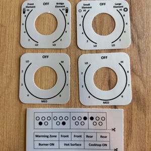 May include: A set of white adhesive control panel overlays for a GE #JS905SK4SS stove. The set includes four square labels with circular cutouts for knobs, featuring temperature markings from LO to HI, and a rectangular sheet with indicator icons.